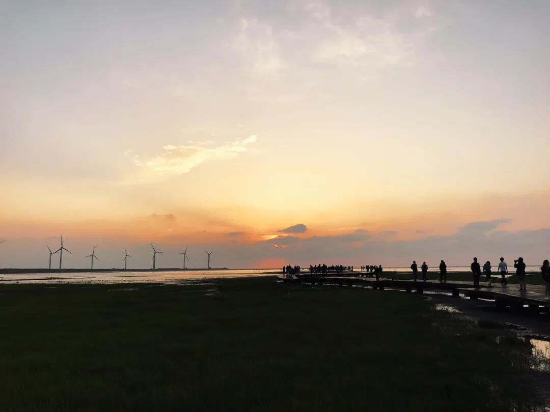 Visitors enjoy the serene beauty of a Gaomei Wetlands sunset from the wooden boardwalk, reflecting the vibrant orange sky.